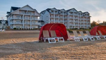 The image shows a beach area with rows of lounge chairs and red canopy tents in front of two large, multi-story buildings.