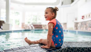 A child in a heart-patterned swimsuit sits by an indoor pool, splashing water and smiling joyfully.