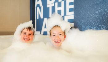 Two children are playing and smiling in a bathtub filled with bubbles, with part of a sign reading 