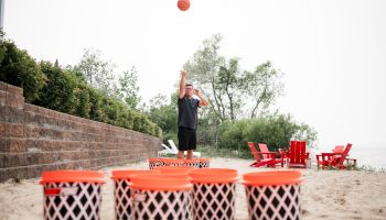 A person on a sandy area is shooting a basketball towards several orange buckets. Red chairs and green bushes are visible in the background.