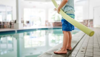 A child stands by an indoor pool holding a yellow pool noodle, wearing swim trunks and a light shirt.