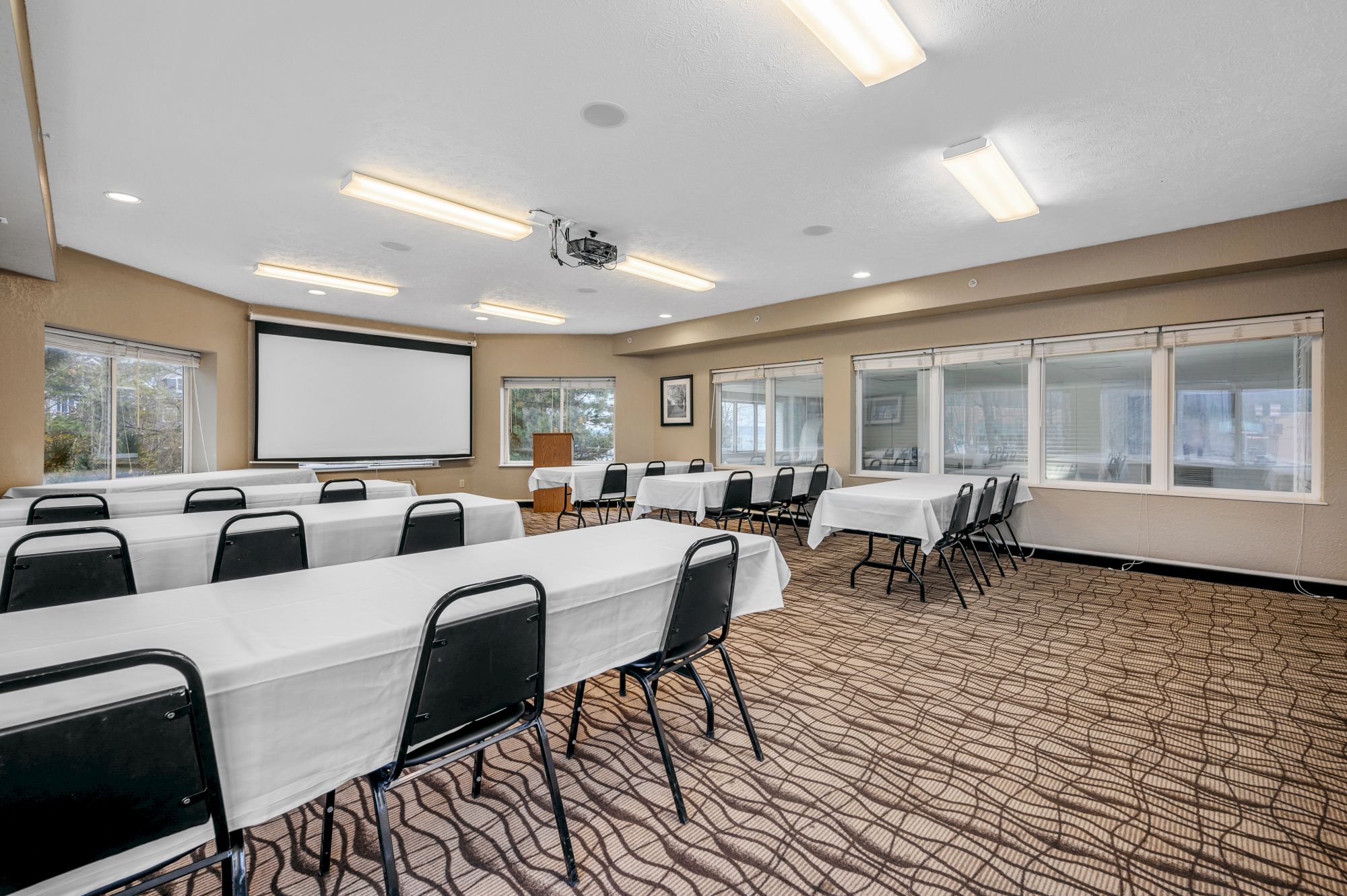 The image shows a conference room with tables and chairs arranged in rows, a projector screen, and windows on two walls.