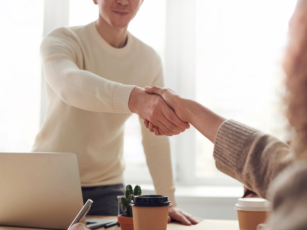 Two people are shaking hands in an indoor setting, with a laptop and coffee cups on the table, in a professional or casual meeting.