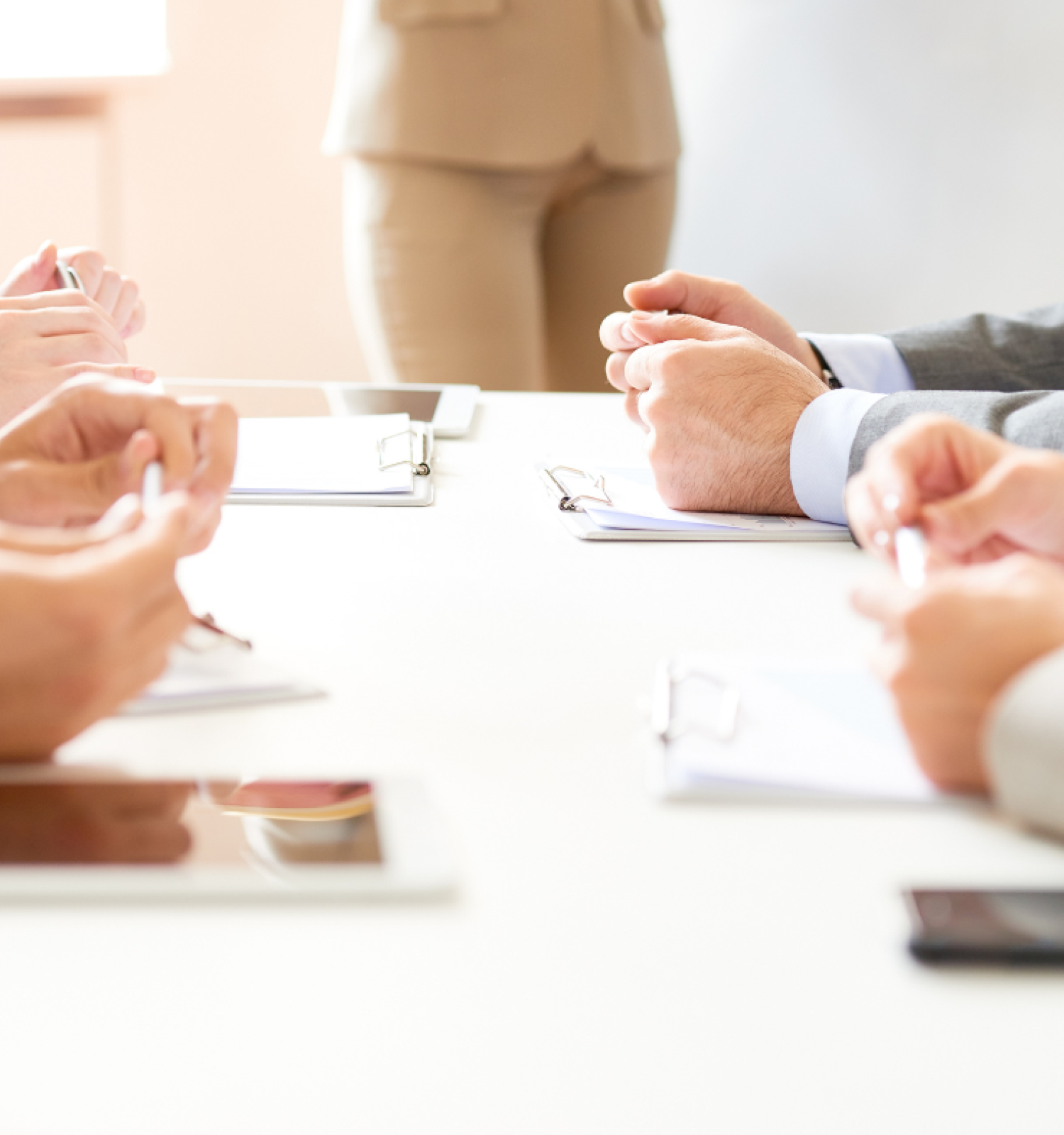 A group of people is seated at a conference table, holding documents and writing on notepads, with a standing person in the background.