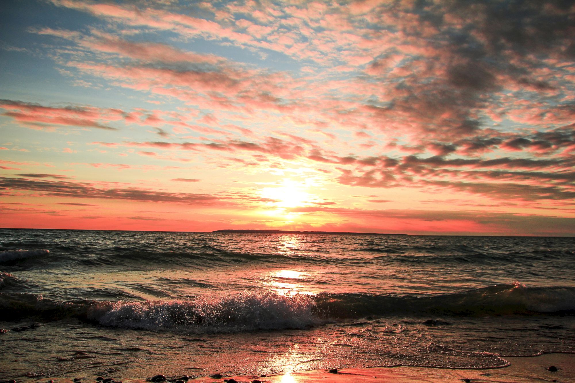 A serene beach scene at sunset with waves gently crashing onto the shore and the sky painted in hues of orange, pink, and blue.