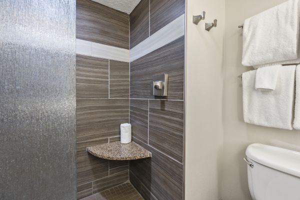 The image shows a modern shower area with grey tiles, a toilet, and towels hanging on the wall. There is also a small granite shelf with tissue paper.