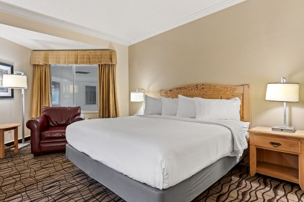 A hotel room with a king-sized bed, white linens, two nightstands with lamps, a brown armchair, a window with curtains, and a small side table.