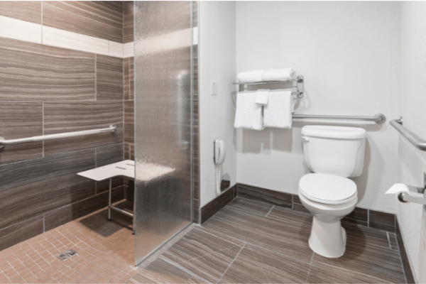 An accessible bathroom with a walk-in shower, grab bars, a wall-mounted foldable shower seat, a toilet, and a towel rack with white towels.