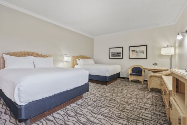 A hotel room with two double beds, a desk with a chair, an additional chair, two framed pictures on the wall, and lamps providing lighting.