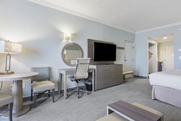 The image shows a modern hotel room with a bed, desk, chair, flat-screen TV, round table with chairs, and minimalistic furnishings, in neutral tones.