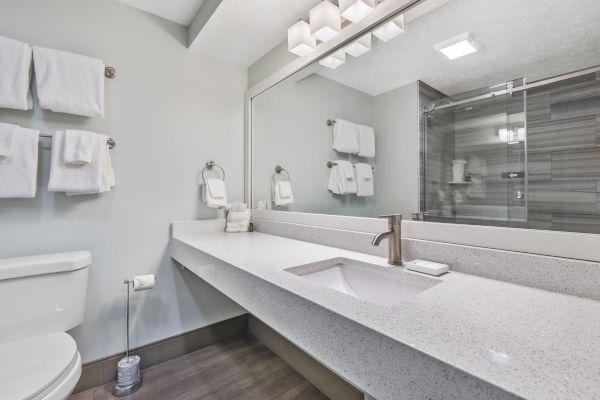 A modern bathroom with a large mirror, white countertop, sink, toilet, and towels on racks. A glass-enclosed shower is visible in the background.