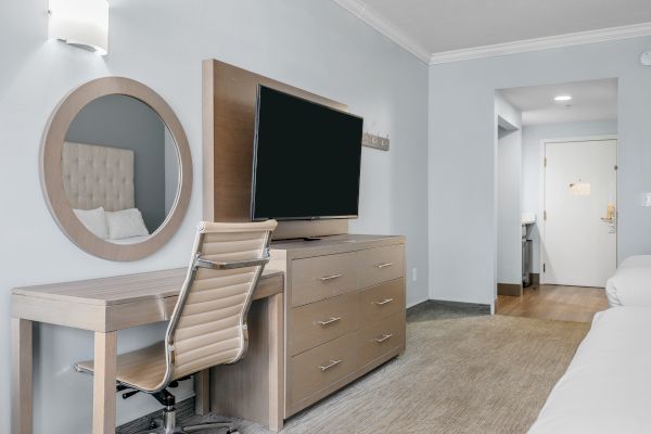 The image features a modern hotel room with a desk, a chair, a large mirror, a flat-screen TV on a dresser, and two beds.