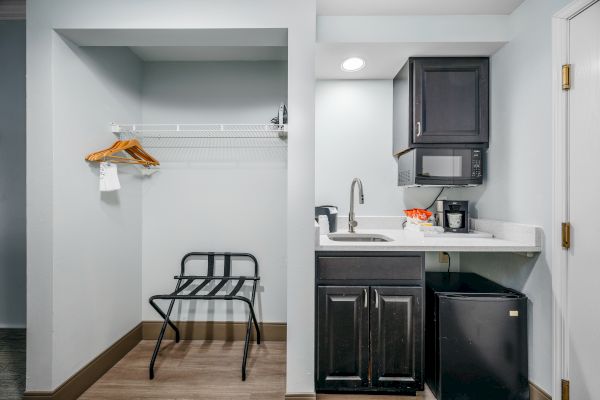 The image shows a small kitchenette with a sink, microwave, coffee maker, mini-fridge, storage cabinets, and a closet area with hangers and a luggage rack.