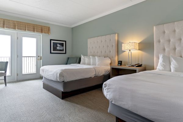 The image shows a modern hotel room with two double beds, a nightstand with a lamp, a chair, and doors leading to a balcony.