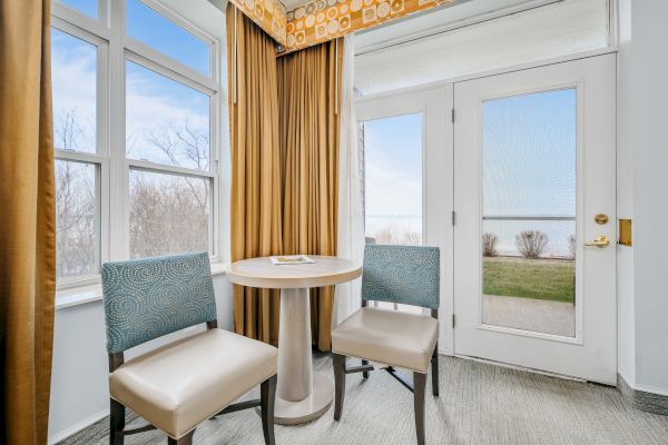 A small seating area with two chairs and a table by a set of windows and a door with a view of the outdoors, including trees and a body of water.