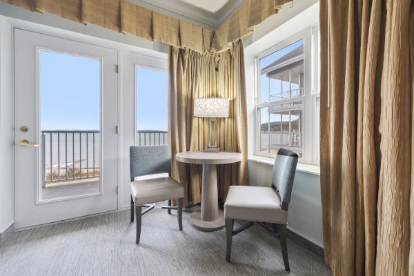 A small, cozy seating area with two chairs and a table next to large windows and a door leading to a balcony, with beige curtains and ample natural light.