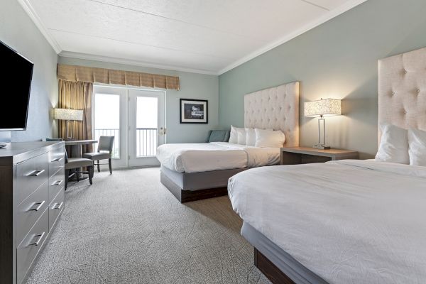 A hotel room with two beds, a TV on the left, a work desk near the window, and a door leading to a balcony.