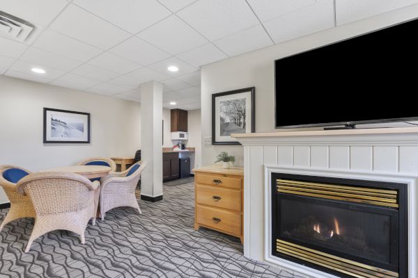 The image shows a cozy living area with a TV above a fireplace, wicker chairs around a table, and a small kitchen area in the background.