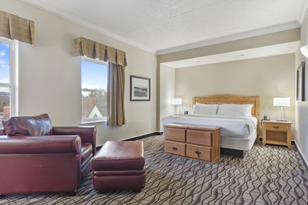 The image shows a spacious hotel room with a king-sized bed, armchair, ottoman, nightstands, and two windows with curtains.