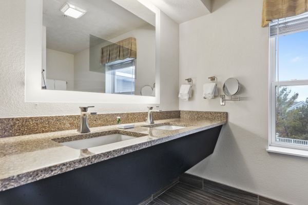 The image shows a modern bathroom with a large mirror, double sinks with sleek faucets, towel holders, and a window allowing natural light.