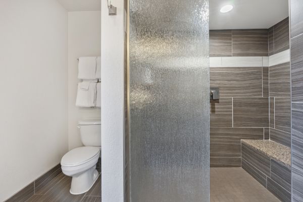 This image shows a modern bathroom with a toilet, towel rack, and a glass-enclosed shower area with grey tiles and a built-in bench.