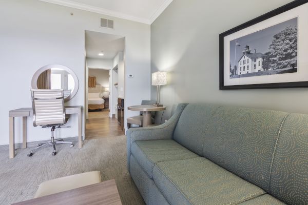 This image shows a living area with a green couch, a photograph on the wall, a desk with a chair, a small dining table, and a visible bedroom.