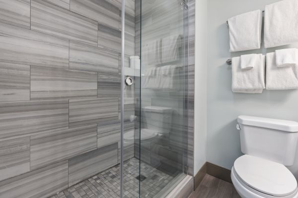 The image shows a modern bathroom with a glass-enclosed shower, gray tile walls, a white toilet, and white towels hung on the wall.