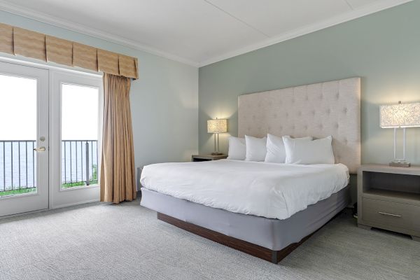 A serene bedroom featuring a large bed with white linens, two bedside tables with lamps, and a door leading to a balcony with a sea view.