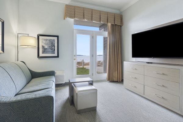 A modern, clean living room with a sofa, TV, and dresser, featuring a door and a window with a view of an outdoor scene.