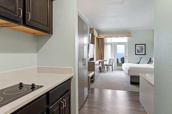 A hotel room with a kitchenette, bed, desk, TV, and a view of the balcony with chairs through a large window.