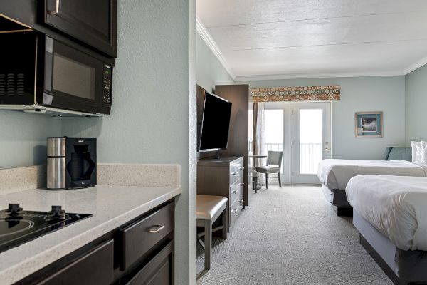 A hotel room with a kitchenette, TV, two beds, and a balcony door. The kitchenette has a stove, microwave, and coffee maker.