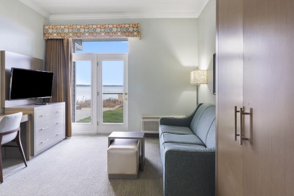 A room featuring a sofa, TV, desk, and chair with a view of a body of water through a french door, and a lamp on the wall.