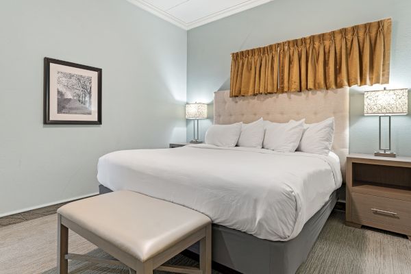 A neatly made bed with white linens, bedside tables with lamps, a framed picture, and a cushioned bench at the foot of the bed.