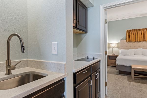 The image shows a kitchenette with a sink and stove, leading into a bedroom with a neatly made bed and a bedside table.