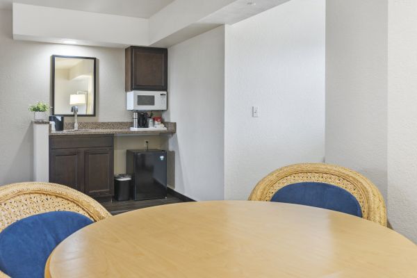 The image shows a small kitchenette with a microwave, mini fridge, and sink, next to a round wooden dining table with four wicker chairs.