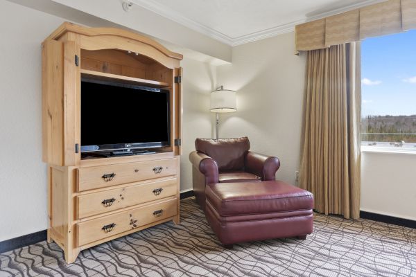 A cozy room featuring a wooden TV cabinet, a leather armchair with an ottoman, a floor lamp, and a window with beige curtains.