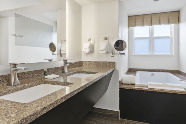 A modern bathroom with double sinks, a large mirror, wall-mounted towels, and a bathtub next to a window, featuring a minimalist design.