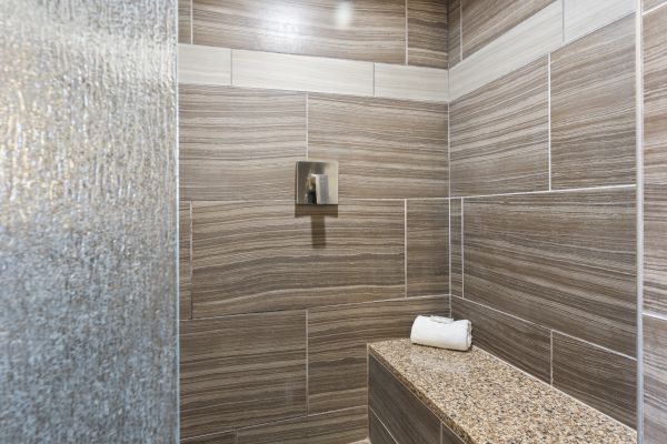 The image shows a modern bathroom shower area with brown and beige tiles, a metallic faucet, and a granite bench with a rolled white towel on top.