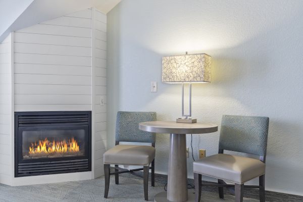 A cozy living area with a fireplace, a round table, two chairs, and a lamp on the table against a light-colored wall.