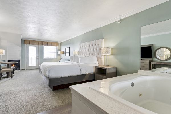 A modern hotel room featuring two beds, a hot tub, a large mirror, a desk, and a TV. The room has soft lighting and a view through French doors.