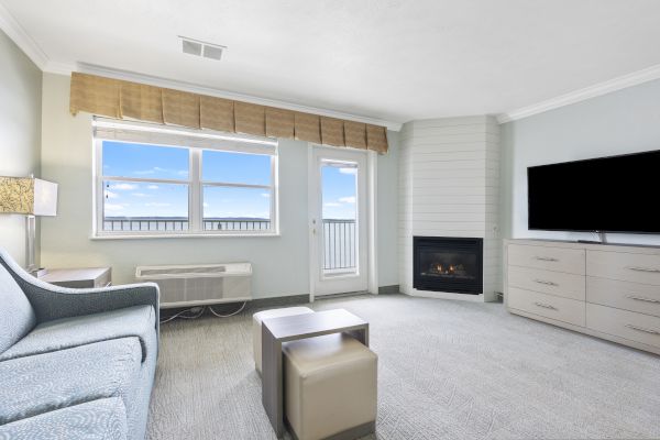 A cozy living room with a sofa, ottoman, TV on a dresser, fireplace, window, and door leading to a balcony with a scenic view of blue sky and clouds.