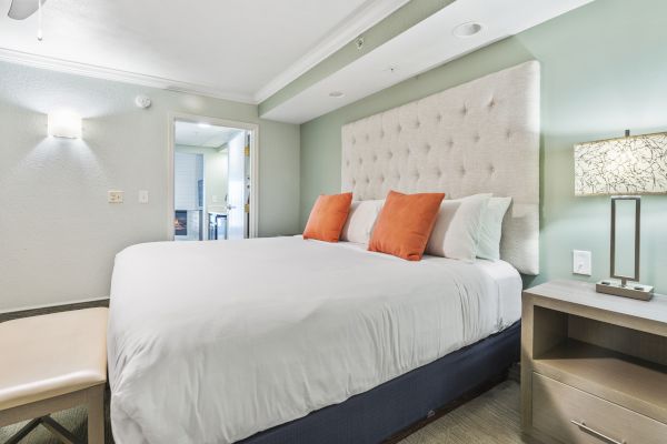 A modern bedroom with a large bed, white linens, orange pillows, a cushioned headboard, a bench, and a nightstand with a lamp.