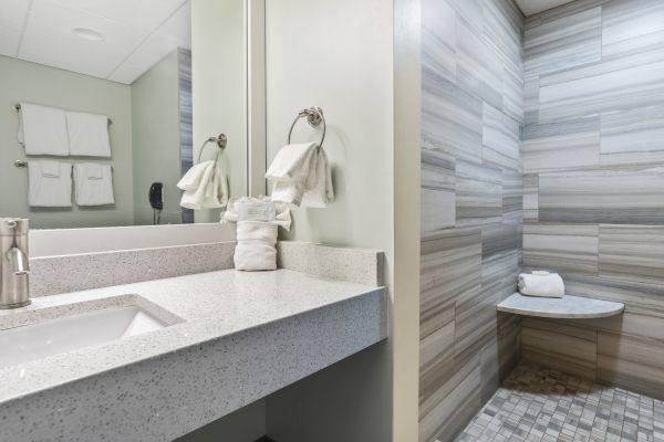 A modern bathroom featuring a sink with a silver faucet and towel, a mirror, and a tiled shower with a bench and white towels.