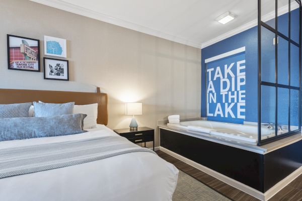 A modern bedroom with a comfortable bed, wall art, a lamp on a nightstand, and an in-room bath area with a blue wall reading "TAKE A DIP IN THE LAKE."