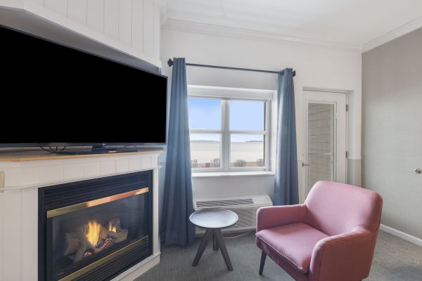 A cozy room with a fireplace, TV, pink chair, small table, and a window with blue curtains overlooking an outdoor view.