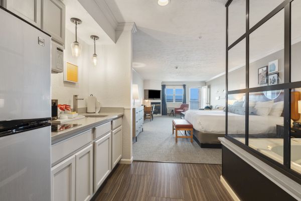 The image shows a spacious hotel room featuring a kitchenette, a large bed, modern furnishings, and a sitting area with windows in the background.