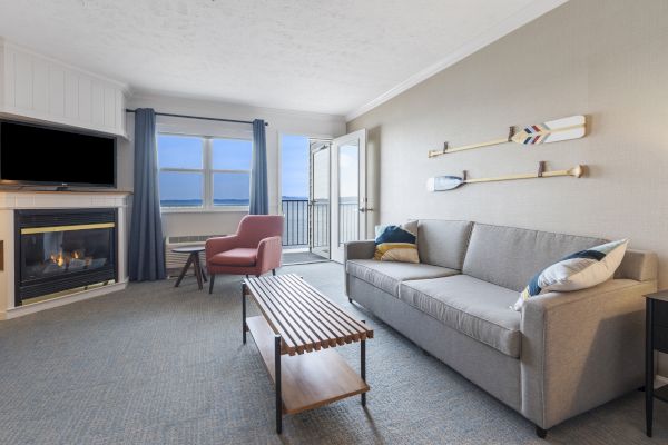 A cozy living room with a fireplace, couch, coffee table, armchair, and nautical decor, overlooking a balcony with an ocean view.