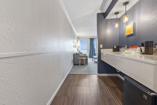 The image shows a hotel room with a hallway leading to a seating area. There’s a mini fridge, countertop, and lights in the foreground.