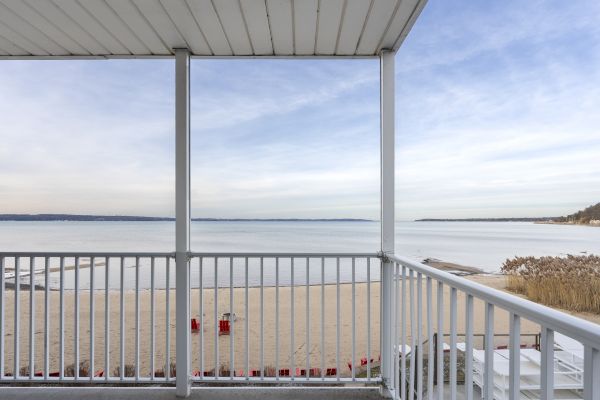 A balcony view overlooking a calm beach and vast ocean under a partly cloudy sky. The scene includes a white railing and a covered roof.