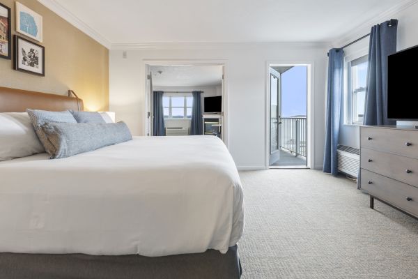 A well-lit bedroom with a large bed, dresser, TV, and two curtained windows. There's also a door leading to an outside balcony.